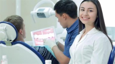 Yeni iş günü dişçileri. Dişçinin yanındaki kız stajyer. Diş hekimi tabletteki beyaz dişli hastayı gösteriyor. Dişleri gülümsüyor ve başparmağını kaldırıyor.