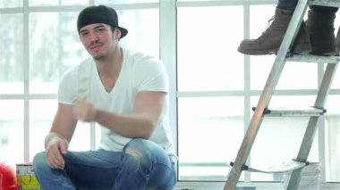 Taşıma ve onarır dairede. Bir pencere pervazına üzerinde oturan ve bir fırça ile boya boş bir dairede bir merdiven üzerinde arka planda duran arkadaşı süre tutarak çalışan ressam gülümseyerek