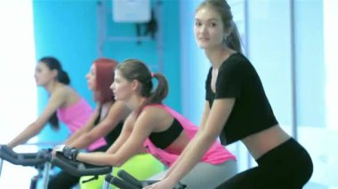 Spor olarak çalışma. Bisiklet arka planda pedal ve sabit bir bisiklet sırasında onun kız arkadaşı sporcular spor salonunda kameraya bakıp atletik kız oldu