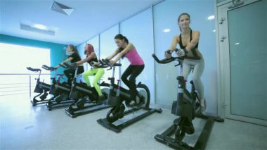 Spor salonunda grup egzersizleri. Pedal arkadaşlar spor ve spor salonunda bir sabit bisiklet önünde görünüyor. Sporcular spor kıyafetler giymiş.