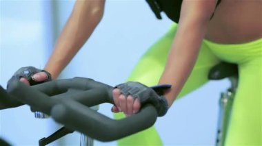 Spor salonunda fitness dersleri. Atletik kız üzerinde simülatörü kadar onun dört kız sporcular spor sabit bir bisiklet üzerinde pedal pedal. Ellerin simülatörü ve spor karın üzerinde kapat