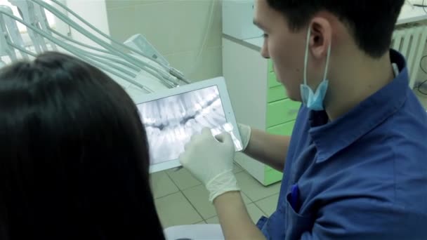 フロントには歯医者 歯科用の椅子の少女 歯科医はタブレットの患者の 線を示し彼女の歯かま補正方法を指示 — ストック動画