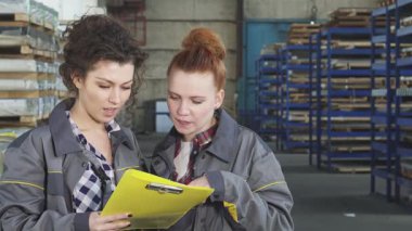 Odaklanmış iki genç bayan depo çalışanı hareketli bir fabrika ortamında gerekli evrakları incelerken canlı bir tartışma içindeler. Ekip çalışmaları organize olarak yol alırken parlıyor.
