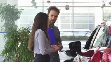 Kendini işine adamış bir araba satıcısı, genç bir bayana parlak ve modern bir galeride seçeneklerini değerlendirirken sıcak bir şekilde yardımcı oluyor. Olgun satıcı, parlak kırmızı bir otomobilin kapısını açar ve onu deneyime davet eder.