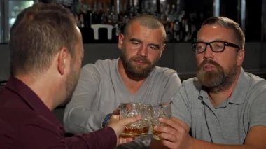 Neşeli, olgun bir adam, arkadaşlarıyla barda keyifli bir gece geçirirken viski bardağını kaldırırken kameraya ışık saçıyor. Sıcak ortam ve kahkaha neşeli bir atmosfer yaratır.