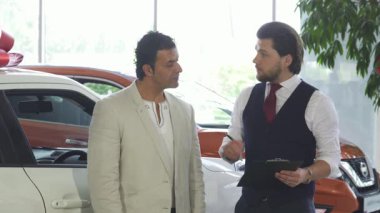 Zarif bir açık renk ceket giyen seçkin olgun bir adam modern bir araba galerisinde bakımlı bir satıcıya evrak imzalatıyor. Parlak sergi salonu, ışıl ışıl