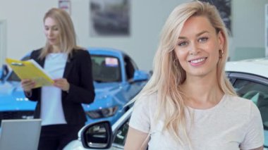 Neşeli bir kadın yeni araba alımını kutlamak için arabasının anahtarlarını gururla tutarken mutluluk saçıyor. Arka planda, profesyonel bir satıcı ona yardım ediyor, canlı bir bayilik sergiliyor.