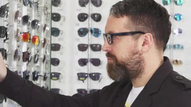 Neşeli sakallı bir adam bir optometriste sergilenen çeşitli şık gözlükleri yakından inceliyor. Heyecan ve merak saçıyor. Moda olan güneş gözlüklerinin göz alıcı dizilimi.