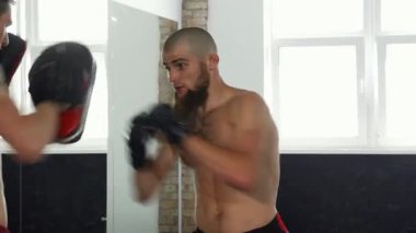 Sakallı bir erkek dövüşçü spor salonundaki yoğun antrenman sırasında pederine güçlü bir yumruk atıyor. Bu heyecan verici sahne takım çalışmasının ve kararlılığın özünü yansıtıyor.