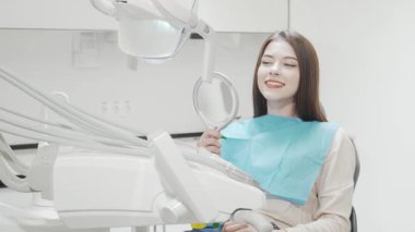 Neşeli genç bir kadın kameraya ışıldıyor, modern bir diş sandalyesinde rahatça oturuyor, parlak gülüşüne hayran olmak için bir ayna tutuyor. Bu canlı sahne diş bakımının özünü yansıtıyor.