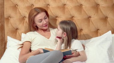 Çekici bir anne ve sevimli küçük kızı, yumuşak yastıklarla çevrili, rahat bir yatakta rahat bir okuma anını paylaşıyorlar. Bu iç açıcı sahne evde eğitimin zevkini yansıtıyor.