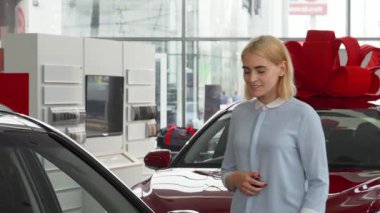 Işıl ışıl genç bir kadın, canlı kırmızı bir yayla süslenmiş çarpıcı yeni otomobilinin anahtarlarını elinde tutarken neşe dolu, gösterişli bir galeride duruyor. Bu an heyecanı yakalar