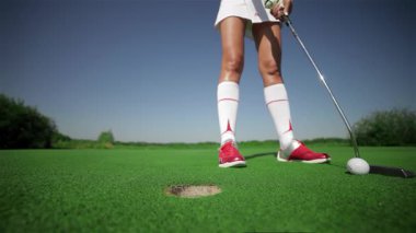 Yakın çekim, golf topuna vurmaya hazırlanan kararlı beyaz bir kadını canlı yeşil bir sahada yakalıyor. Şık, beyaz bir etek ve tişört giymiş olarak atletizmini sergiliyor.