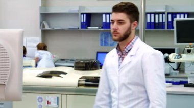 Beyaz laboratuvar önlüklü genç beyaz bir adam yüksek teknoloji laboratuarındaki parlak bir monitöre doğru emin adımlarla ilerliyor. Düşünceli ifadesi ve bakımlı sakalı bilimsel çalışmalarına olan bağlılığını yansıtıyor.