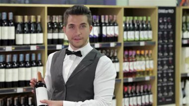 Şık bir smokin giymiş genç ve çekici bir şarap garsonu, şarap şişesini düzgünce düzenlenmiş şarap raflarının önünde gururla sunar. Kendinden emin gülüşü ve özgüvenli duruşu profesyonelliği gözler önüne seriyor.