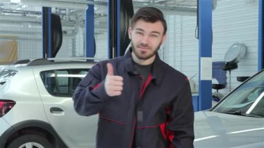 Becerikli sakallı bir tamirci gösterişli bir araba servisinde dikilirken başparmağını kaldırır. Siyah ve kırmızı üniformalı bu genç beyaz adam, kollarıyla profesyonellik sergiliyor.