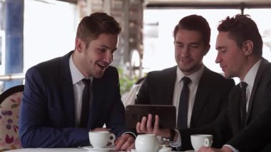 Şık bir kafede, üç iyi giyimli adam bir tableti incelerken dikkatle planlarını tartışıyor. Atmosfer canlı ve profesyonel, dumanı tüten bardaklarla...