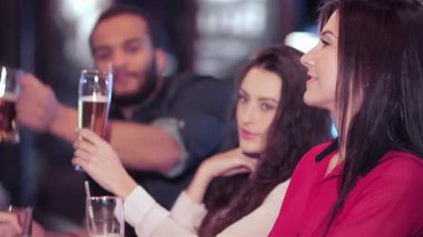 Canlı bir grup genç arkadaş, hem erkek hem de kadın, hareketli bir bar atmosferinde bira bardaklarıyla neşeyle kadeh kaldırıyorlar. En sevdikleri futbol takımı için tezahürat yapmanın heyecanı havayı dolduruyor.