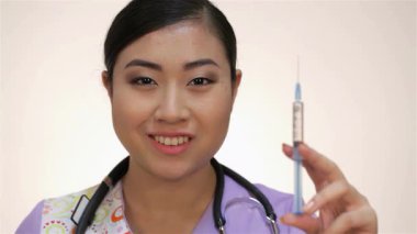Kendine güvenen genç bir Asyalı kadın doktor, beyaz bir elbise giymiş, yumuşak beyaz bir arka plana karşı dostane bir gülümsemeyle bir şırınga tutuyor. Stetoskop gibi tıbbi aletlerle çevrili.