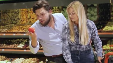 Çekici bir çift canlı domatesleri hareketli bir süpermarkette keşfediyor. Sakallı beyaz adam olgun kırmızı domatesi inceliyor. Çekici sarışın kadın gülümsüyor ve domates ekliyor.