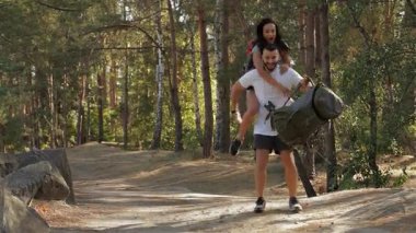 Neşeli bir bayan yürüyüşçü, canlı bir orman zemininde erkek arkadaşlarının üzerine sevinçle atlıyor. Yakışıklı sakallı adam, neşeli bir gülümseme ile, sırt çantası tutarken zahmetsizce onu destekliyor.