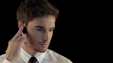 Gıcır gıcır beyaz gömlekli ve parlak siyah kravatlı ciddi bir beyaz adam, dramatik bir siyah arka plan karşısında, kulaklığından dikkatle dinliyor. Düşünceli ifadesi ve ince hareketleri.