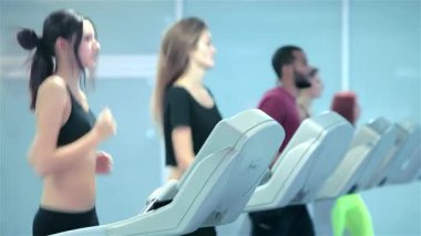 Hem erkek hem de kadın sporcular da dahil olmak üzere bir grup kendini adamış sporcu koşu bantlarında enerjik bir şekilde koşuyor ve spora olan bağlılıklarını sergiliyorlar. Şık spor kıyafetleri içinde, renkli vücutları yansıtıyor.