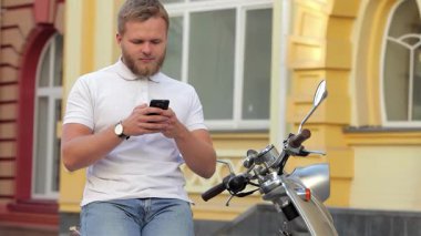 Sakallı şık bir genç adam şık scooterına biner, akıllı telefonuyla canlı bir şehir arka planına takılır. Bu canlı sahne, yaz özgürlüğünün ve modern hayatın özünü yansıtıyor.