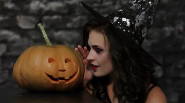 Büyüleyici bir kadın büyücü, sırıtan bir balkabağının yanına eğilir, parmağını kaldırır ve karanlık, desenli tuğla bir duvara karşı susturur. Bu Cadılar Bayramı esinlenilmiş sahne gizem saçıyor