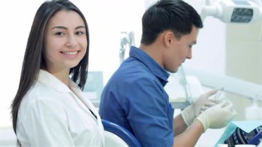 Neşeli bir diş asistanı, dişçi aletlerini özenle hazırlayan odaklı bir dişçinin yanında otururken parlak bir gülümseme yayar. Bu ilgi çekici sahne modern bir takım çalışmasının özünü yakalar.