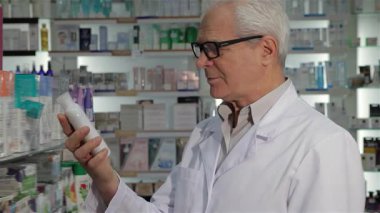 Gıcır gıcır beyaz laboratuvar önlüğü giymiş yaşlı bir erkek eczacı elinde bir şişe ilaç tutarken nazikçe gözlüğünü çıkarıyor. Onun dostane tavrı ve canlı ilaç ürünleri rafları.