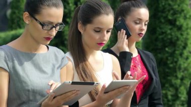 Üç kendine güvenen iş kadını şık resmi kıyafetler giyip tabletlerine odaklanırken, biri telefonuyla konuşuyor ve dinamik bir çalışma ortamı sergiliyor. Özgüvenli ifadeleri ve