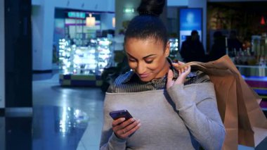 Şık bir Afrikalı Amerikalı kadın, hareketli bir alışveriş merkezinde canlı alışveriş torbalarıyla çevrili akıllı telefonuyla neşe içinde sohbet ediyor. Onun ışıltılı gülümsemesi ve günlük kıyafetleri eğlenceli bir alışveriş deneyimini yansıtıyor.
