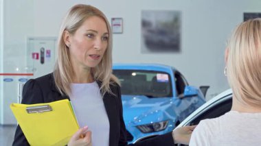 Kendinden emin bir satıcı kadın, parlak otomobillerle çevrili şık bir araba galerisinde müşterisiyle seçenekleri tutkuyla tartışır. Atmosfer profesyonellik ve heyecanla uğulduyor.