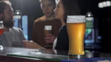 Güzelce hazırlanmış bir bira bardağı tezgahın üzerinde belirgin bir şekilde dururken, bir grup canlı arkadaş barda kutlama için bardaklarını tokuşturuyor. Bar ışıklarının sıcaklığı şenliği arttırır.