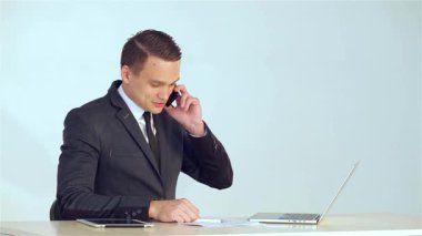 Parlak takım elbiseli gülümseyen genç bir işadamı aktif olarak bir telefon görüşmesi yapıyor, profesyonellik ve karizma yayıyor. Masasında bir dizüstü bilgisayar ve tabletle modern şirketin özünü somutlaştırıyor.