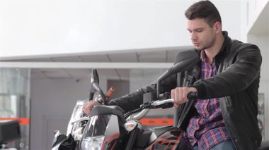 Hafif sakallı, genç, beyaz bir adam gösterişli, siyah bir motosikletin benzin deposuna yaslanıyor. Kendine güveni ve tarzı tam. Ekose gömleği ve deri ceketi modern olanı tamamlıyor.