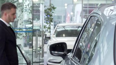 Sivri takım elbiseli genç bir işadamı, modern bir galeride şık bir arabayı inceliyor. Düşünceli ifadesi ve etrafındaki parıldayan araçlar bir heyecan ortamı yaratıyor.