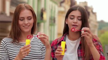 İki güzel genç kadın, biri uzun kahverengi saçlı ve diğeri dalgalı koyu saçlı, neşeli bir şehir caddesinde canlı sabun köpükleri üflüyor. Kahkahaları havayı dolduruyor.