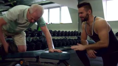 Kendini adamış genç beyaz bir antrenör, üst düzey bir müşterinin spor salonunda egzersiz yapmasını dikkatle izliyor ve destekleyici bir atmosfer sergiliyor. Sakallı fitness uzmanı yaşlı adamın yanında diz çöker.