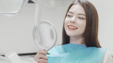 Işıl ışıl bir kadın, dişçi kliniğindeki aynada parlak gülüşünü incelerken sağlıklı dişlerini sergiliyor. Bu canlandırıcı sahne diş bakımının zevkini yakalar,