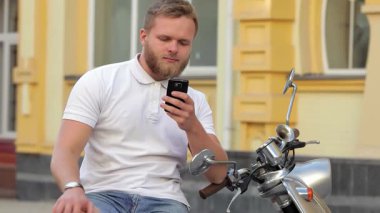 Şık sakallı neşeli bir genç adam scooterına binmiş, akıllı telefonuna dalmış. Canlı kentsel zemin, özgürlük ve macera hissini artırıyor ve bu sahneyi mükemmel kılıyor.