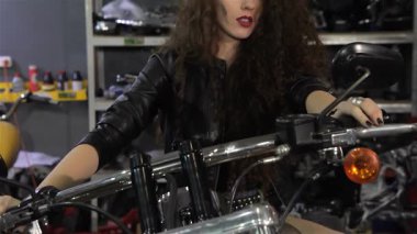 Büyüleyici genç bir bayan motorcu özgüven ve cazibeyle motorsiklet tankına doğru incelikle eğiliyor. Kıvırcık saçlarıyla yüzünü çerçeveliyor, baştan çıkarıcı bir şekilde kameraya bakıyor, şık bir şekilde giyinmiş.