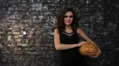 Büyüleyici bir kadın büyücü, sırıtan bir balkabağını tutarken baş parmağını kaldıran siyah tuğlalı bir duvara karşı güven içinde poz veriyor. Bu Cadılar Bayramı 'ndan esinlenilmiş görüntü neşe saçıyor.