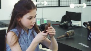 Endişeli küçük bir kız, uzun ve hasarlı saçlarını güzellik aletleriyle çevrili şık bir salon ortamında inceler. İfade dolu yüzü, saçla baş etmenin duygusal mücadelesini yakalıyor.