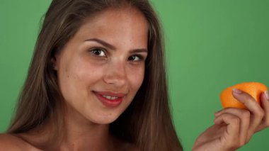 Canlı bir stüdyo çekimi, canlı yeşil bir krom arka planda sulu bir portakalı tutarken kameraya neşeyle gülümseyen parlak derili genç bir kadını yakalar. Bu resim pozitifliği somutlaştırır
