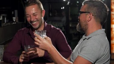 İki arkadaş, sakallı, olgun bir adam ve şık yoldaşı, canlı bir bar ortamında viski bardaklarını tokuştururken bir anlık kahkaha atarlar. Bu canlı sahne özünü yakalar.