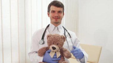 Parlak, hoş geldin ofisinde, koyu saçlı ve sakallı bir erkek doktor sevimli bir oyuncak ayıyı tutarken ışıldıyor. Beyaz laboratuvar önlüğü ve mavi eldivenler giymişti. Büyük bir hevesle başparmaklarını gösteriyordu.