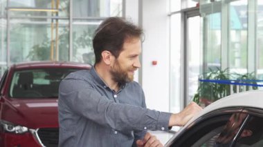 Neşeli, yakışıklı, sakallı bir adam lüks bir galeride yeni arabasının yanında poz verirken neşe saçıyor. Enerjik atmosfer heyecanla dolu, memnuniyetini vurguluyor.