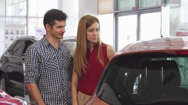 Şık bir çift, canlı kırmızı bluzlu bir kadın ve sıradan gömlekli bir adam, galeride parlak yeni bir otomobili inceliyor. Onların neşeli ifadeleri heyecanı yansıtır ve